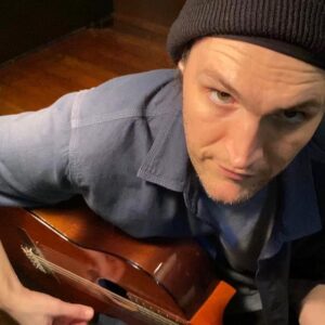 Fotografia de Josh Klinghoffer tirada de um ângulo superior. Josh veste um gorro preto e uma camisa azul, enquanto segura um violão.